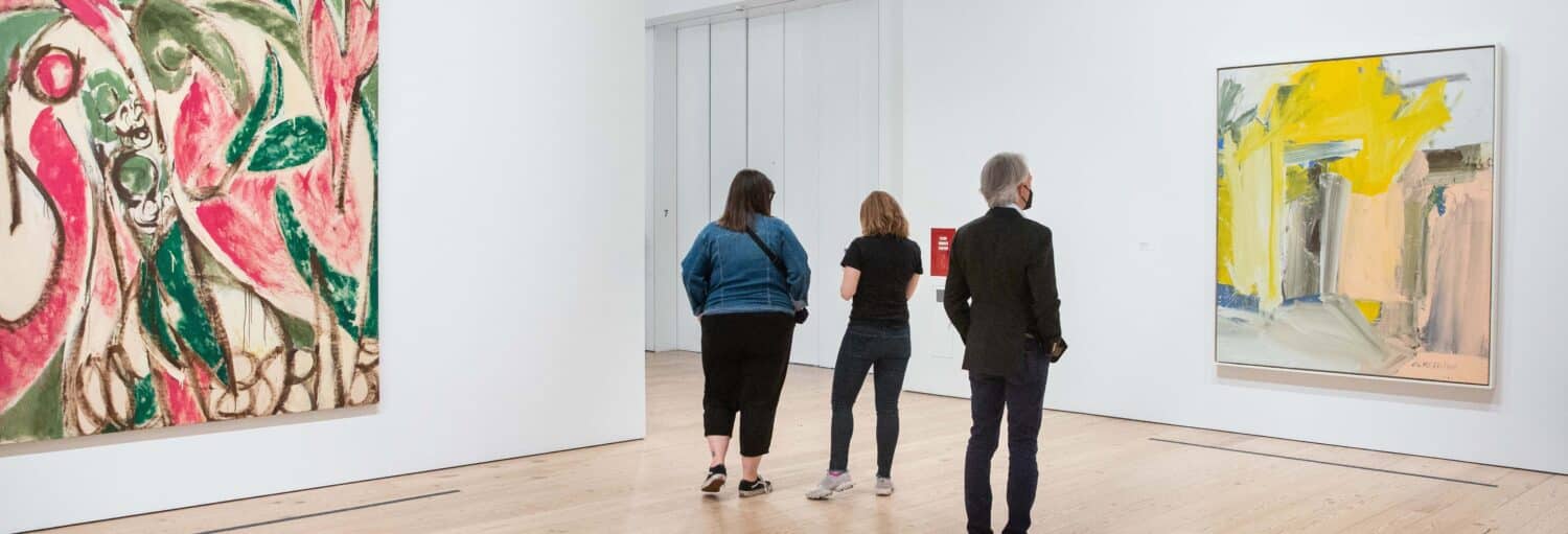 Três pessoas observam pinturas abstratas em uma galeria de arte espaçosa, com paredes brancas e piso de madeira. A foto é capa do post de Ingresso do Whitney Museum of American Art.