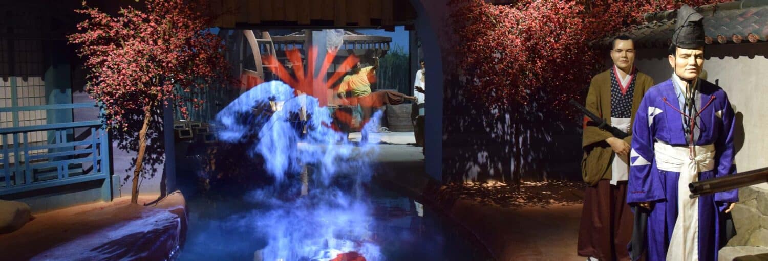 Dois manequins em trajes tradicionais japoneses estão perto de um canal, com um efeito de fumaça azul e árvores vermelhas nas proximidades. A foto é capa do post do Ingresso do World of Discoveries.