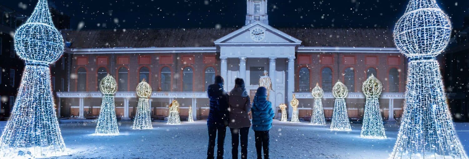 Três pessoas estão do lado de fora de um prédio à noite, cercadas por grandes e brilhantes exibições de luzes natalinas enquanto a neve cai.