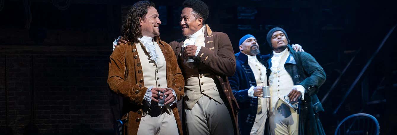 Quatro atores em trajes de época estão no palco, interagindo e sorrindo durante uma apresentação teatral. A foto é capa do post Ingressos para Hamilton.