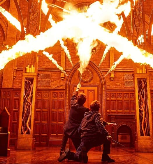Seis pessoas em um palco apontam varinhas, atravessando correntes de fogo em um cenário gótico, semelhante a uma catedral, com iluminação laranja. A foto é capa do post Ingressos para Harry Potter e a Criança Amaldiçoada.