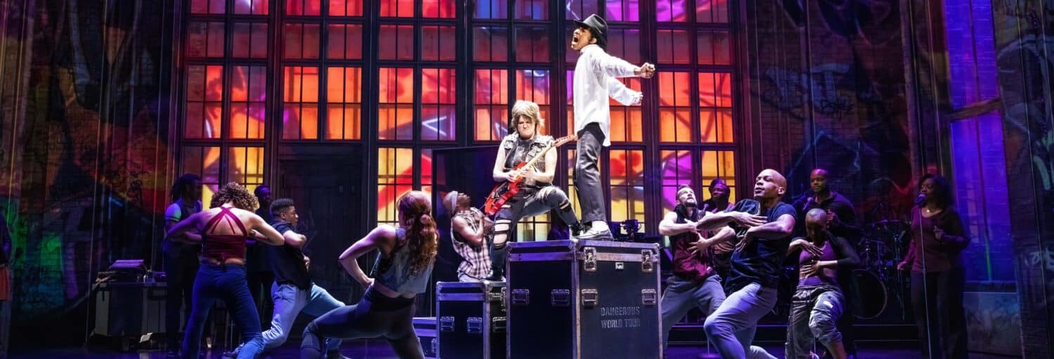 Um grupo de dançarinos se apresenta energicamente no palco com luzes coloridas e duas pessoas em pé em grandes caixas pretas no centro. A foto é capa do post Ingressos do MJ, o musical do Michael Jackson.