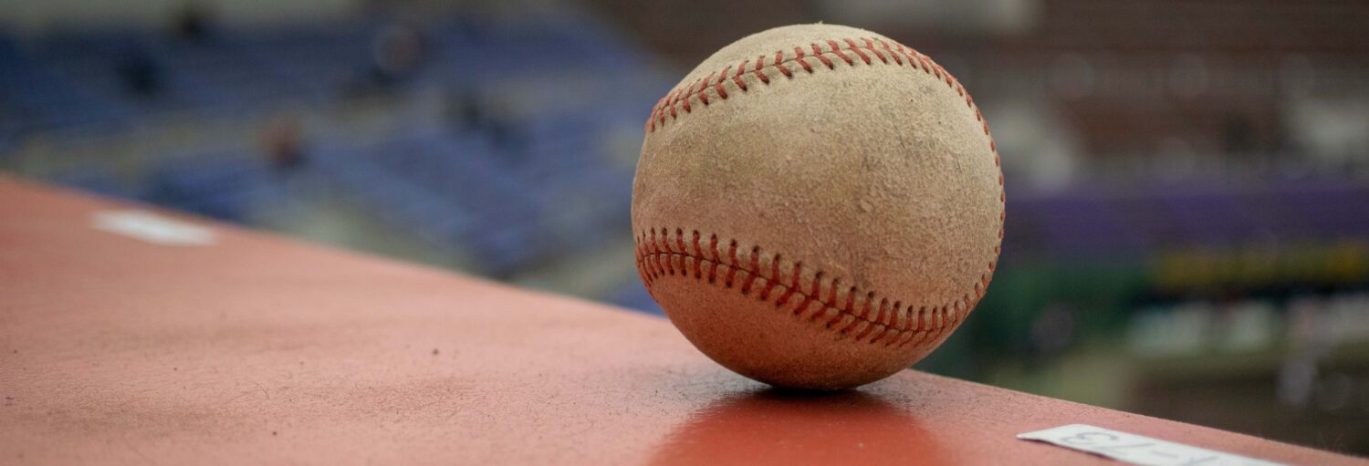 Um close de uma bola de beisebol gasta sobre uma superfície vermelha, com os assentos do estádio desfocados ao fundo.