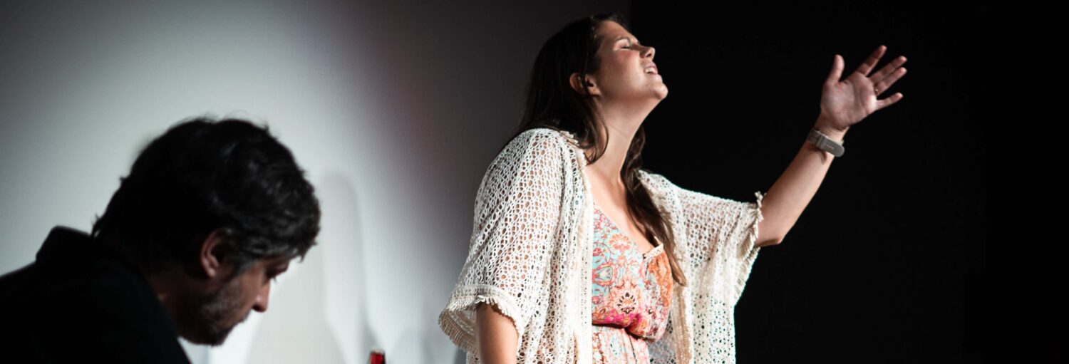 Uma mulher gesticula com o braço levantado enquanto canta; um homem está sentado em primeiro plano com a cabeça baixa. A foto é capa do post do Espetáculo de fado na Baixa.