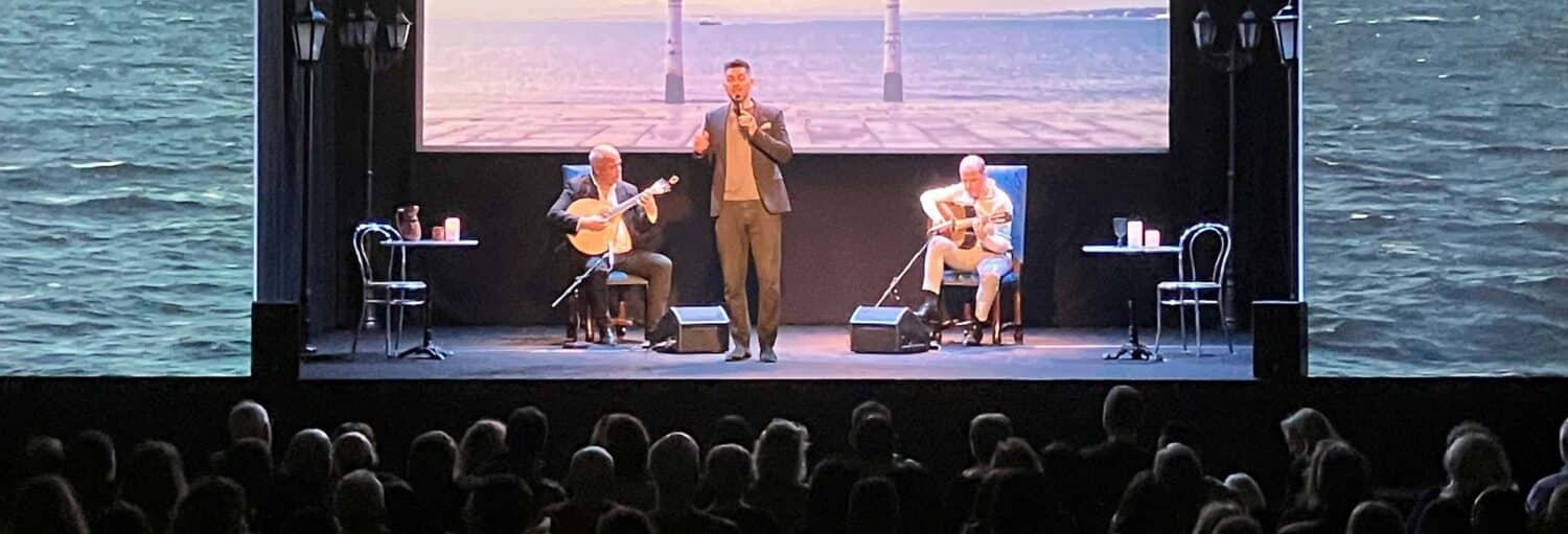 Três artistas estão no palco com guitarras; uma plateia observa enquanto a paisagem oceânica é exibida ao fundo. Imagem para ilustrar o post sobre espetáculo de fado no Chiado.