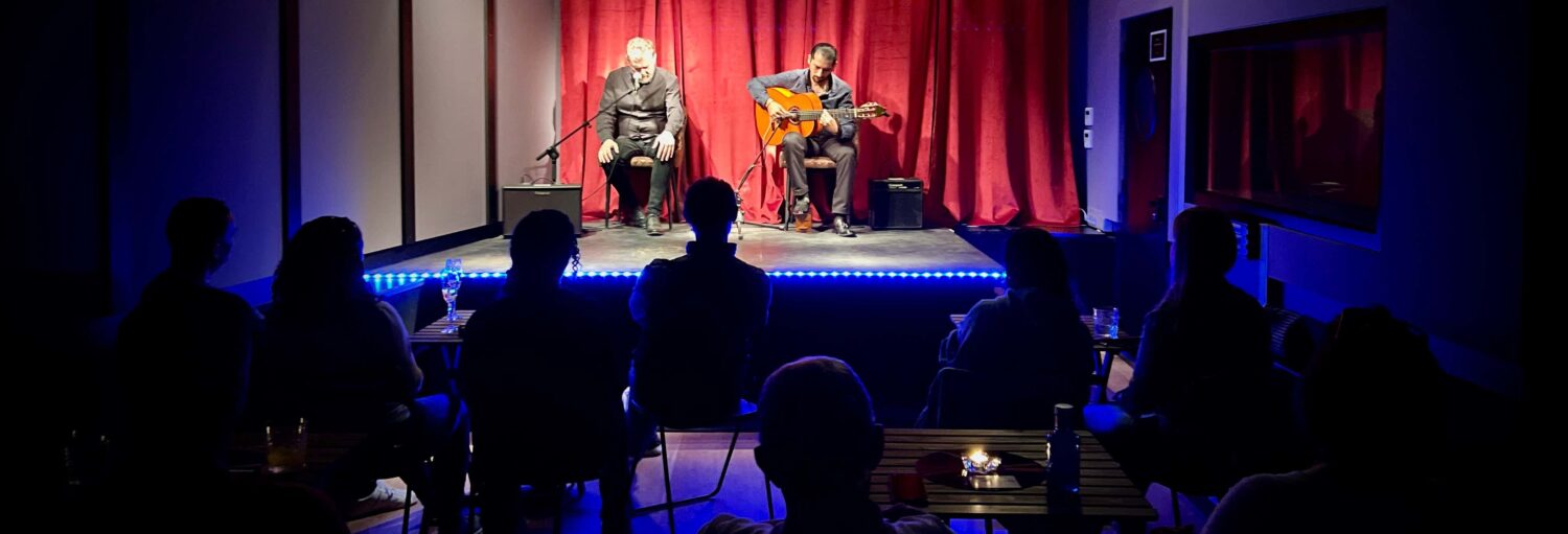 Dois músicos se apresentam no palco com um violão e um microfone para um público sentado em mesas em uma sala pouco iluminada. A imagem ilustra o post sobre Flamenco na Casa Sors + Museu do Violão.