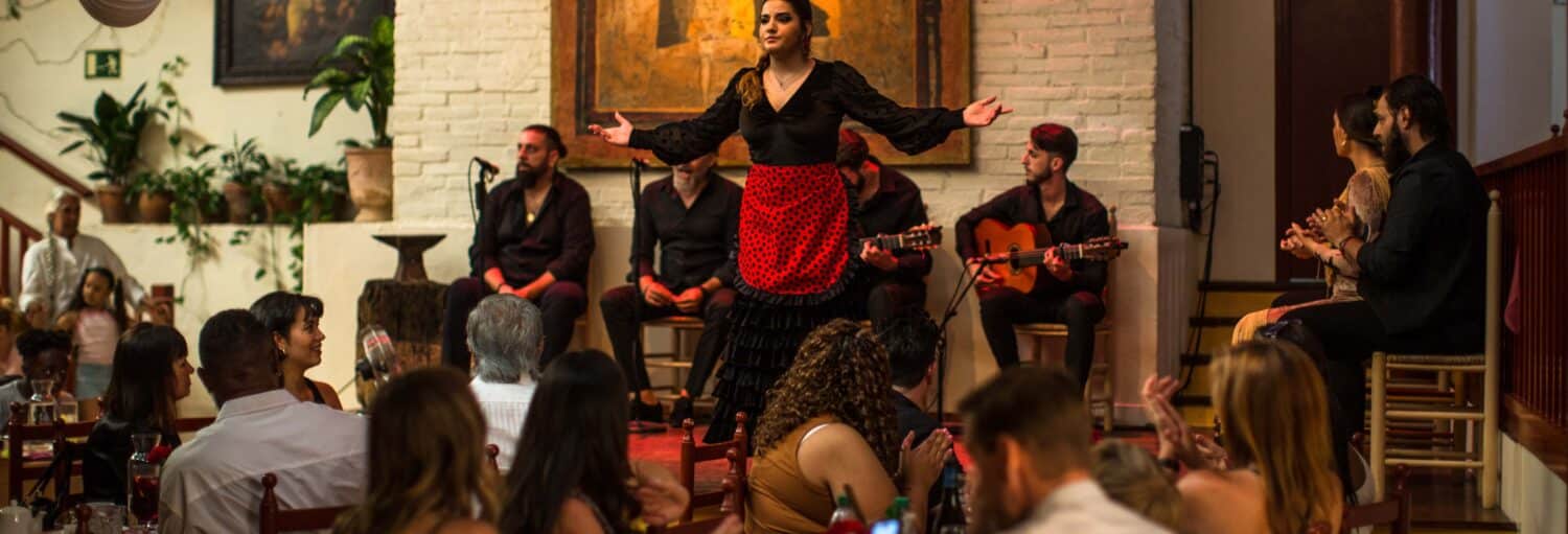 Uma mulher em uma roupa preta e vermelha de flamenco se apresenta no palco com músicos enquanto o público assiste em um restaurante. A imagem ilustra o post sobre espetáculo flamenco no Tablao de Carmen.