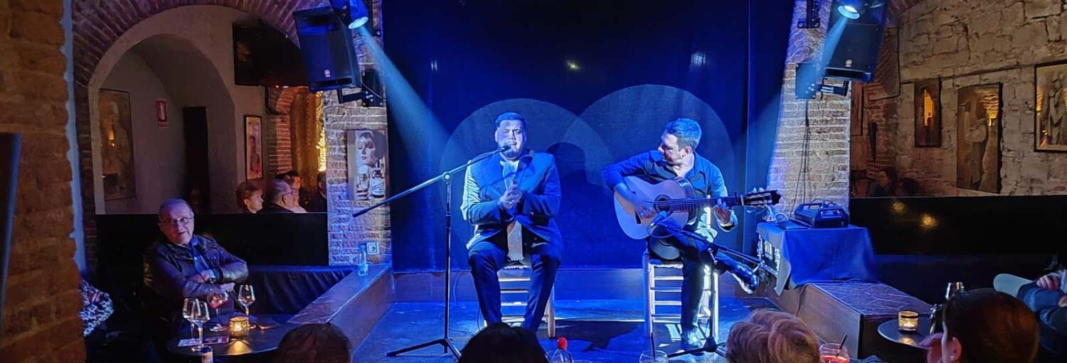 Dois homens se apresentam no palco em um local pouco iluminado; um canta em um microfone, o outro toca um violão. O público assiste. A imagem ilustra o post sobre espetáculo no Tablao Flamenco El Paraigua.