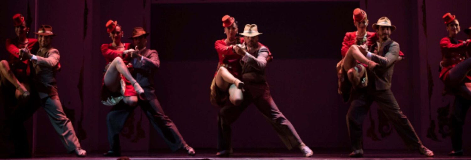 Um grupo de pares em trajes vintage dança no palco sob uma iluminação dramática, cada homem mergulhando seu parceiro. Imagem para ilustrar post sobre espetáculo de tango no Café de los Angelitos.