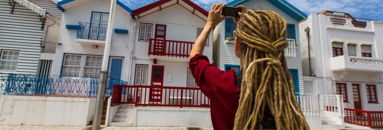 Uma pessoa com longos dreadlocks loiros tira uma foto de casas coloridas e listradas com varandas em um dia ensolarado. A foto é capa do post da Excursão a Aveiro e Costa Nova + Passeio de barco.