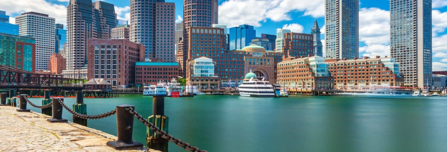 Orla de Boston com edifícios altos e modernos, barcos atracados e águas calmas sob um céu parcialmente nublado. A foto é capa do post Excursão a Boston.