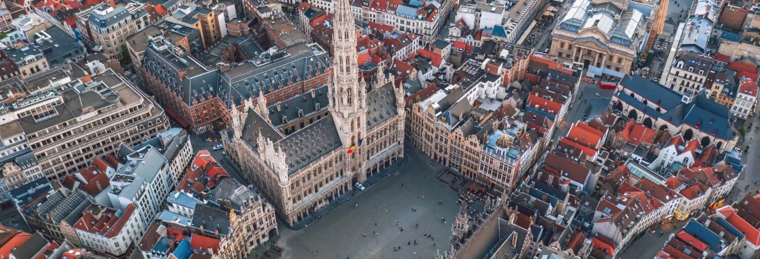 Vista aérea de um grande edifício histórico em uma praça cercada por prédios com telhados vermelhos e ruas estreitas, ilustrando a página da excursão a Bruxelas e Bruges