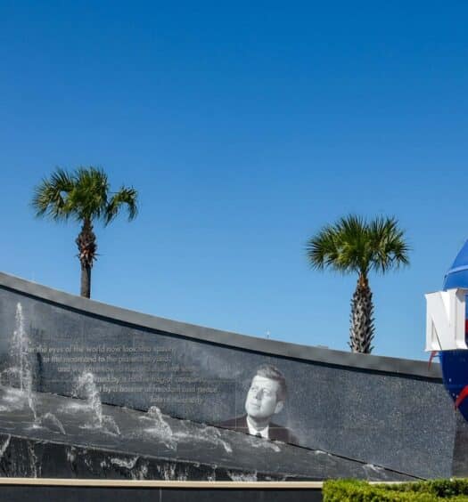 Uma grande escultura de globo da NASA fica ao lado de uma fonte com palmeiras e um foguete ao fundo, sob um céu azul, no Centro Espacial Kennedy.