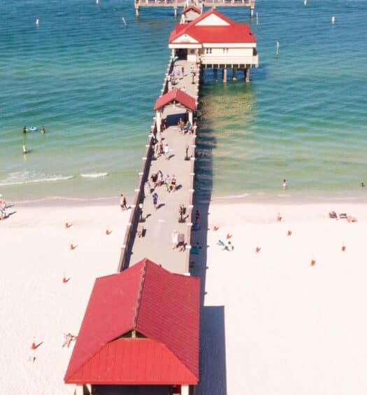 Vista aérea de um píer com abrigo de telhado vermelho estendendo-se sobre o oceano a partir da praia de Clearwater Beach, na Flórida, com areia com pessoas e guarda-sóis.