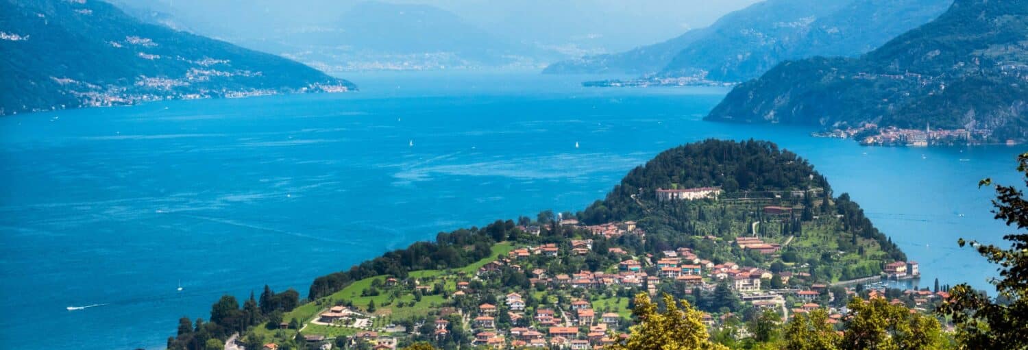 Vista de uma cidade costeira e um lago azul grande com montanhas ao fundo. Excursão a Como e Bellagio + Passeio de barco