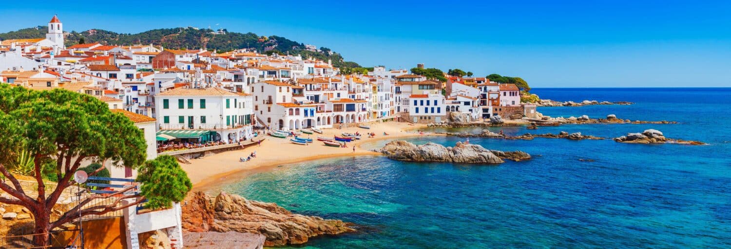 Prédios brancos com telhados vermelhos têm vista para uma praia de areia e mar azul claro em um vilarejo costeiro do Mediterrâneo. A imagem ilustra o post sobre Excursão à Costa Brava.