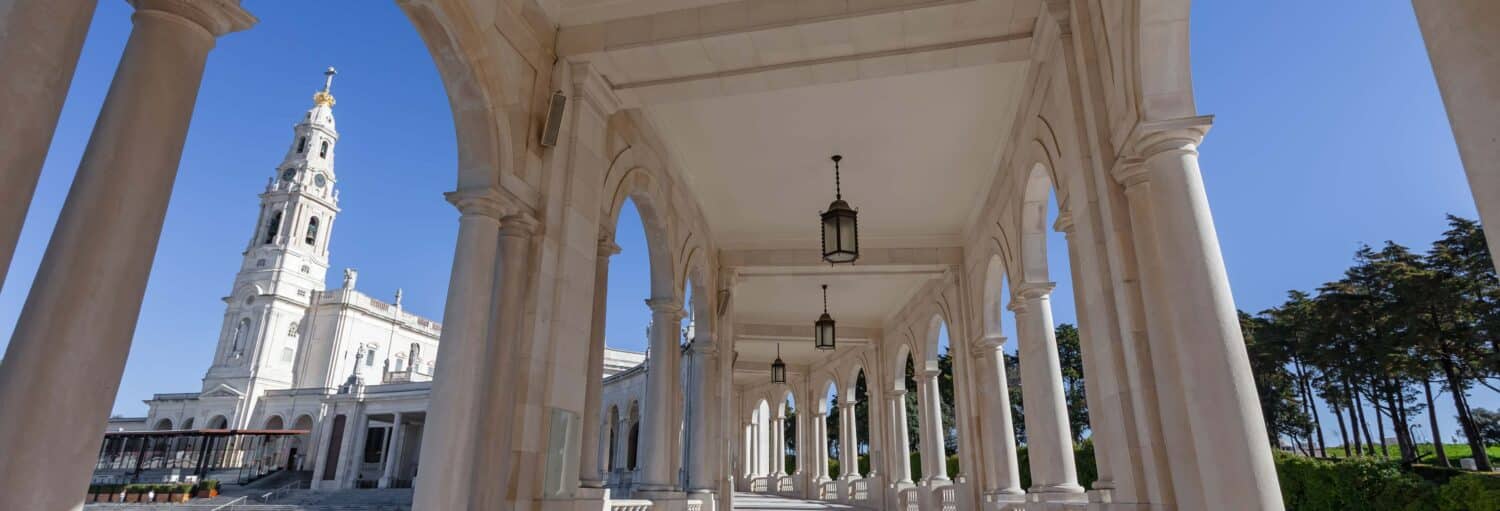 Passarela coberta com arcos de pedra e lanternas penduradas perto da torre de uma igreja branca sob um céu azul claro. A foto é capa do post da Excursão a Fátima e Coimbra.
