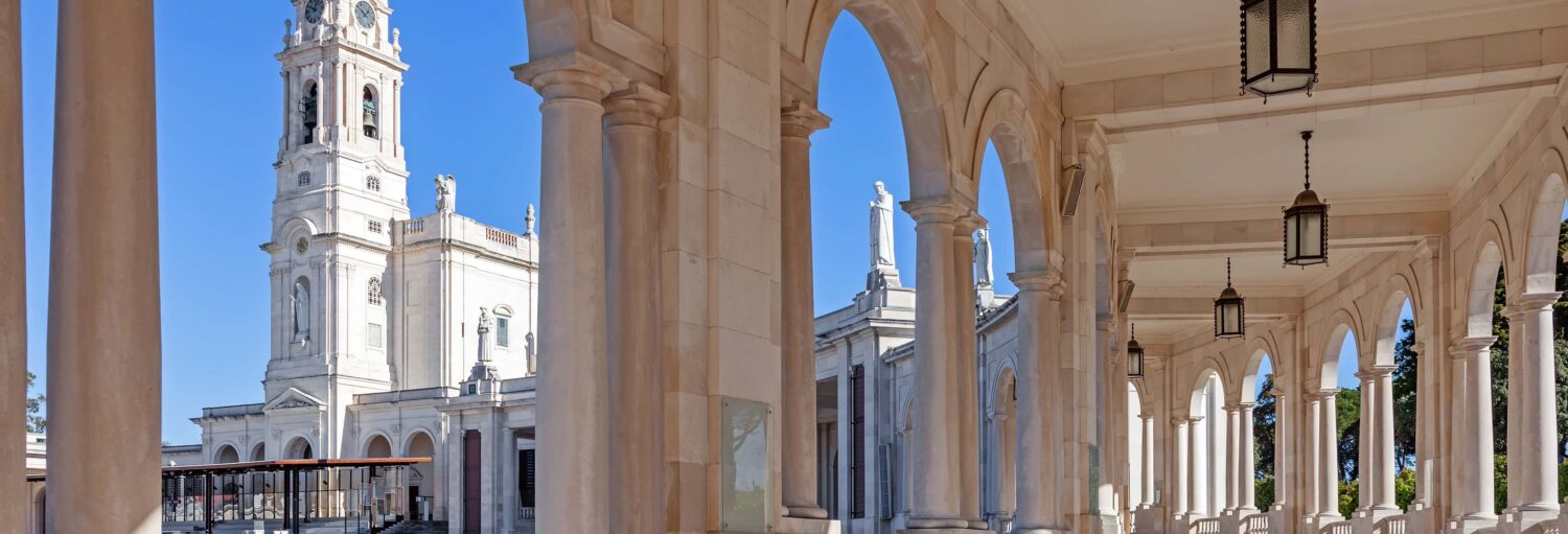 Vista do Santuário de Fátima, em Portugal, mostrando arcos, colunas e a torre da basílica sob um céu azul claro. Imagem para ilustrar post sobre excursão a Fátima e Santarém.
