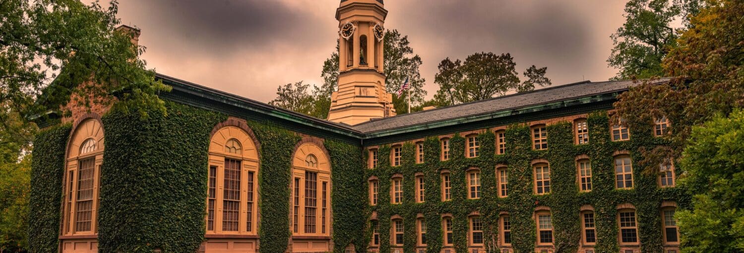 Um edifício histórico coberto de hera com uma cúpula central, grandes janelas e árvores ao fundo sob um céu nublado. A foto é capa do post Excursão à Filadélfia e à Universidade de Princeton.