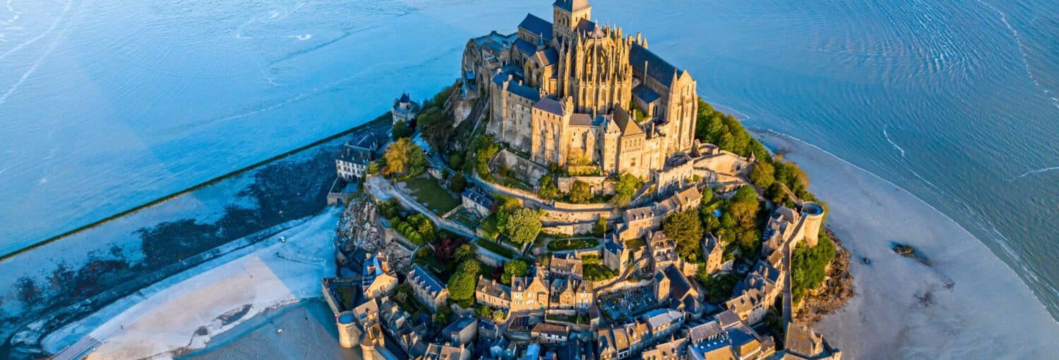 Vista aérea do Monte Saint-Michel, uma comuna histórica com uma abadia, cercada por água na maré baixa. A imagem ilustra o post sobre excursão ao Mont Saint Michel