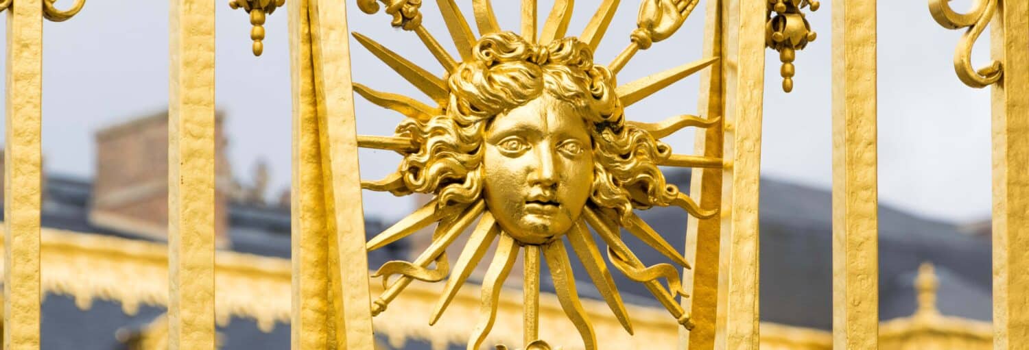 Close-up de um portão dourado com um sol esculpido com um rosto humano e raios radiantes. A imagem ilustra o post sobre excursão ao Palácio de Versalhes de ônibus.