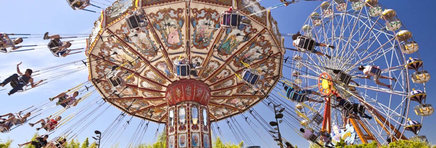 Pessoas andando em um carrossel de balanço colorido e em uma roda-gigante em um parque de diversões em um dia ensolarado. Imagem para ilustrar post sobre Excursão ao Parque de la Costa.