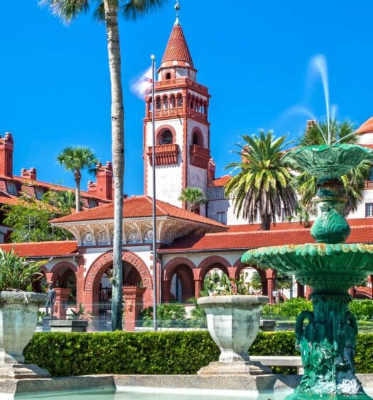Um edifício histórico com telhado vermelho, arcos e torres fica atrás de uma fonte verde e palmeiras em um dia ensolarado durante a excursão a Saint Augustine