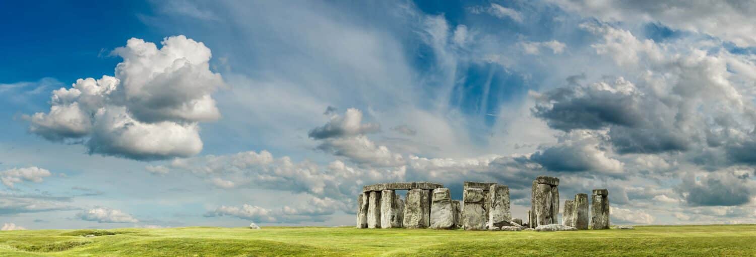 Vista de um campo com várias rochas que formam um círculo. O céu é azul e com nuvens. Excursão a Stonehenge e Bath