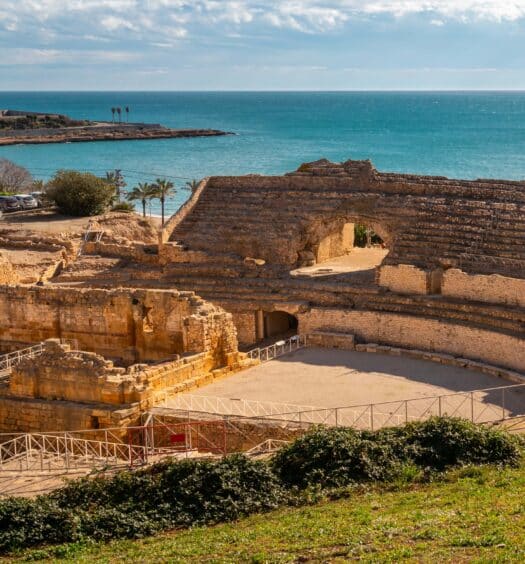 Ruínas de um antigo anfiteatro romano perto da costa com edifícios ao fundo e o mar sob um céu parcialmente nublado. A imagem ilustra o post sobre Excursão a Tarragona e Sitges.