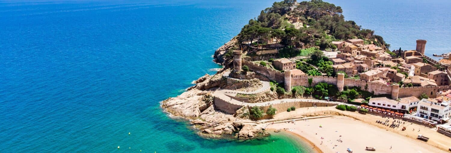 Vista aérea de uma fortaleza costeira com muros de pedra, edifícios e uma praia de areia ao lado de um mar azul claro. A imagem ilustra o post sobre Excursão a Tossa de Mar.