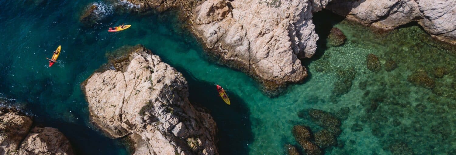 Vista aérea de três caiaques remando em uma água azul-turquesa cristalina cercada por penhascos rochosos e rochas submersas. A imagem ilustra o post sobre Excursão a Tossa de Mar + Mergulho ou caiaque.