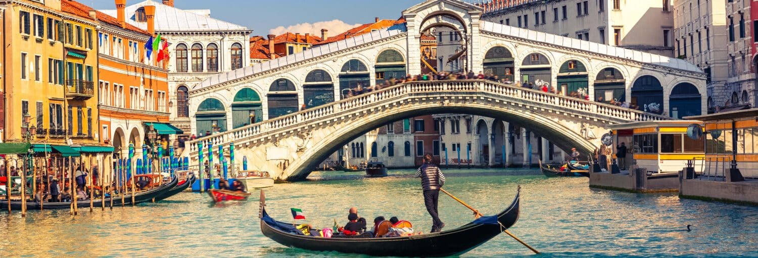 Um barco com pessoas dentro passando por um canal de Veneza. Há uma ponte centralizada mais à frente do barco, e na esquerda é possível ver casas coloridas. Excursão a Veneza