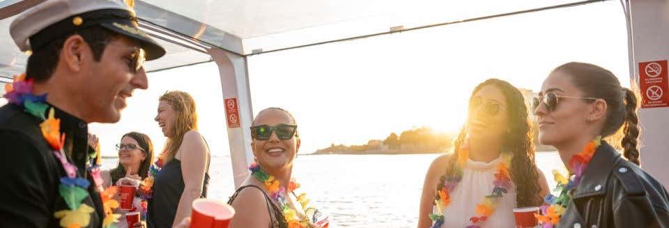 Pessoas usando colares e óculos escuros conversam e sorriem em um barco durante o pôr do sol, segurando copos vermelhos. A foto é capa do post da festa de barco pelo rio Douro.
