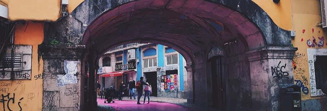 As pessoas caminham e se reúnem sob um arco de pedra em uma rua coberta de grafite, com calçada rosa e prédios coloridos. Imagem para ilustrar post sobre free tour pela Lisboa boêmia.