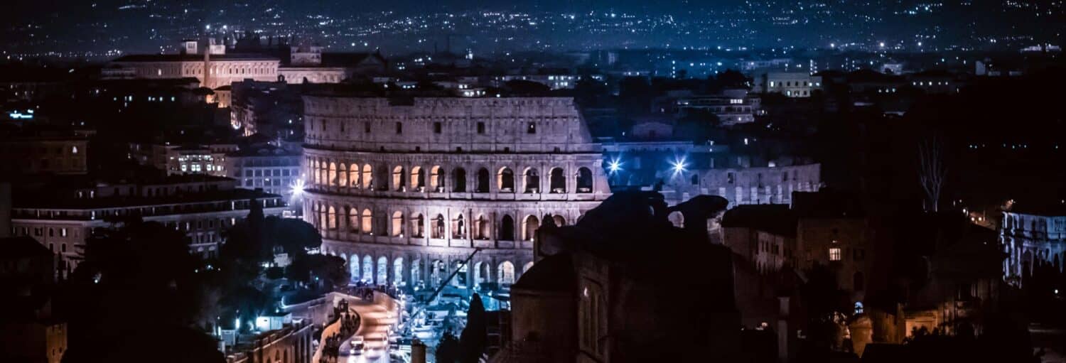 O Coliseu em Roma iluminado à noite, cercado por prédios da cidade e luzes distantes no Free tour de mistérios e lendas por Roma
