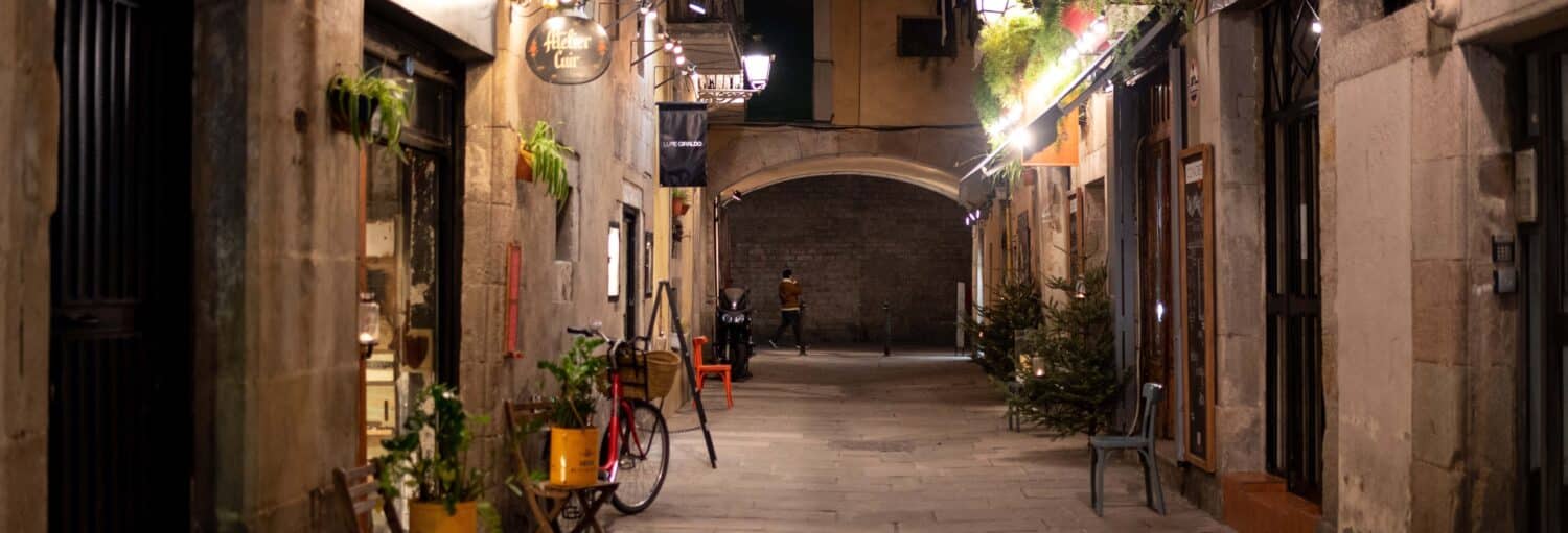 Rua estreita de pedra à noite com lojas, plantas, cadeiras, uma bicicleta e uma pessoa caminhando sob um arco à distância. A imagem ilustra o post sobre um free tour dos mistérios e lendas de El Born.