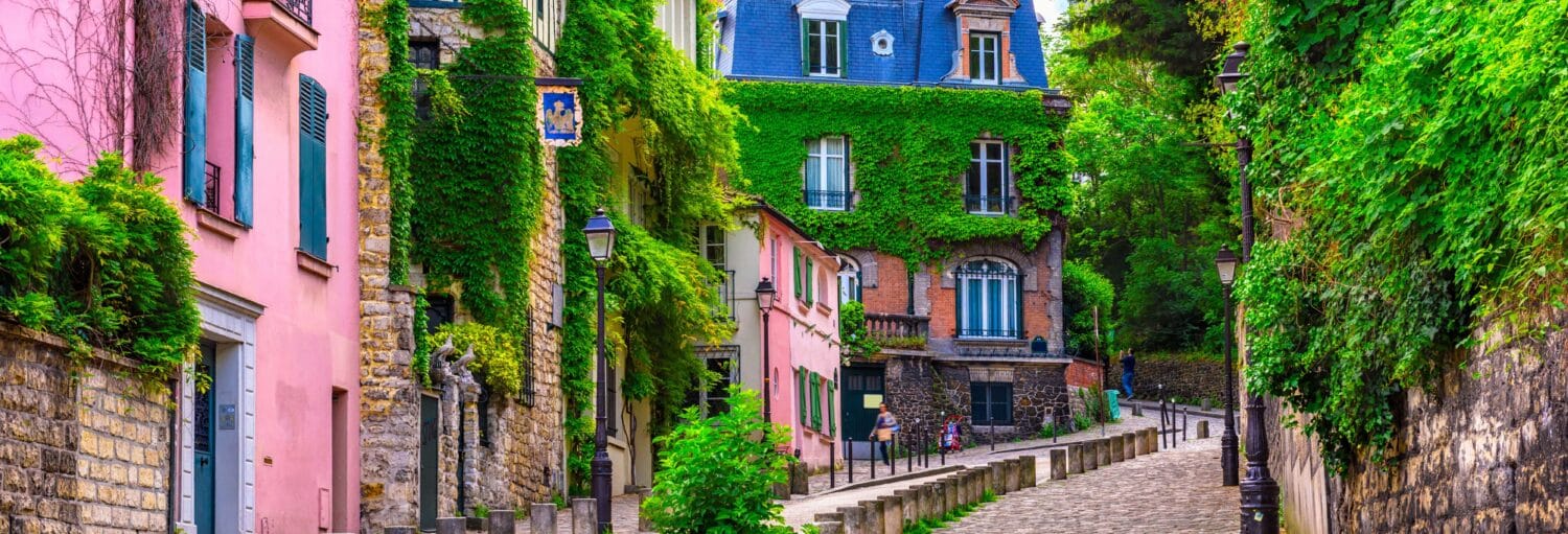 Uma rua de paralelepípedos ladeada por prédios cobertos de hera e muito verde em um tranquilo bairro europeu. A imagem ilustra o post sobre free tour por Montmartre.