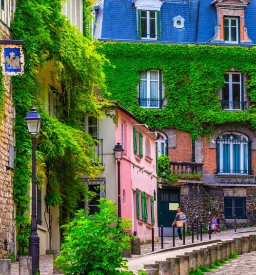 Uma rua de paralelepípedos ladeada por prédios cobertos de hera e muito verde em um tranquilo bairro europeu. A imagem ilustra o post sobre free tour por Montmartre.