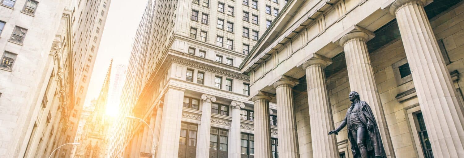 Prédios altos e colunas de pedra com uma estátua em primeiro plano, com a luz do sol brilhando entre os prédios. A foto é capa do post de Free tour por Nova York.
