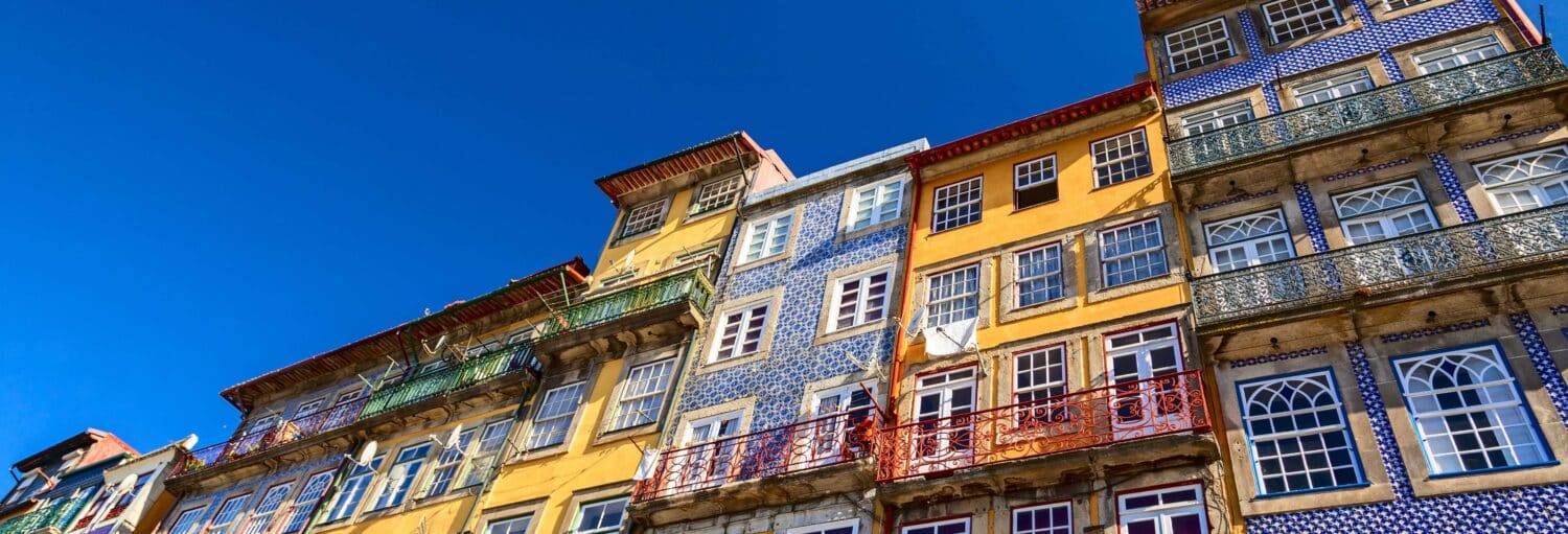 Prédios coloridos com azulejos e varandas padronizados contra um céu azul claro, vistos de um ângulo baixo. A foto é capa do post do Free tour pelo Porto.