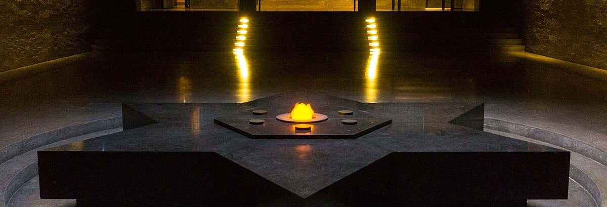 Uma chama queima no centro de um monumento de pedra escura em forma de estrela, em uma sala mal iluminada. A imagem ilustra o post sobre free tour da Segunda Guerra Mundial.