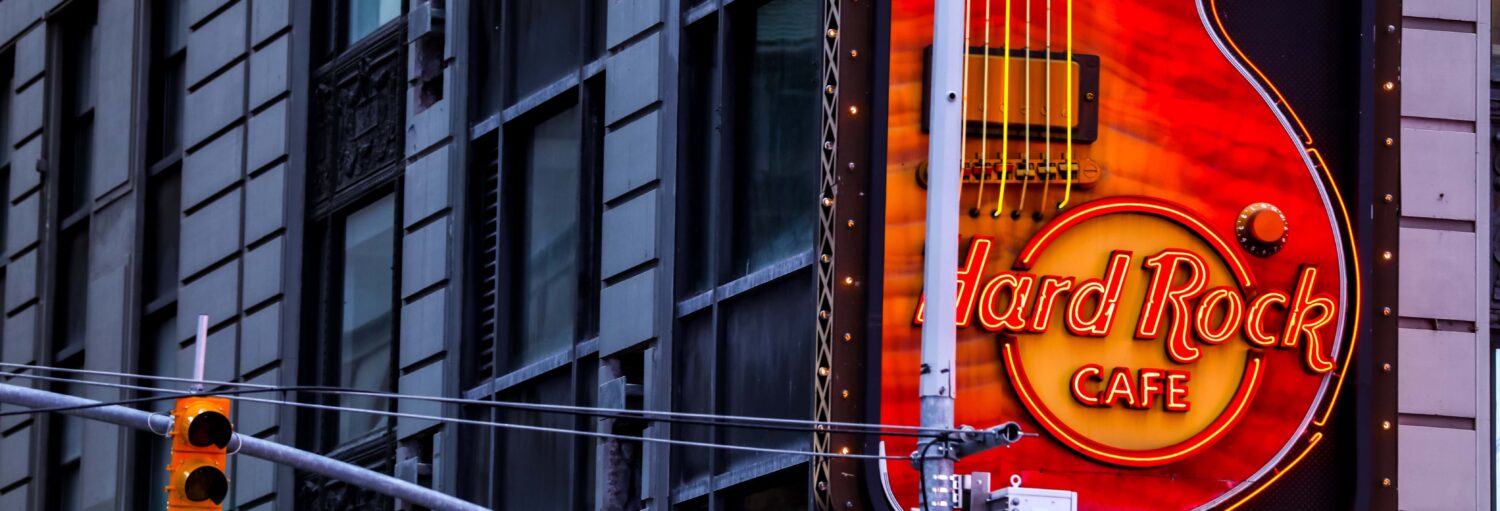 Um letreiro iluminado do Hard Rock Cafe em forma de guitarra está montado na lateral de um prédio da cidade, próximo a um semáforo. A foto é capa do post Hard Rock Cafe Nova York com acesso prioritário.
