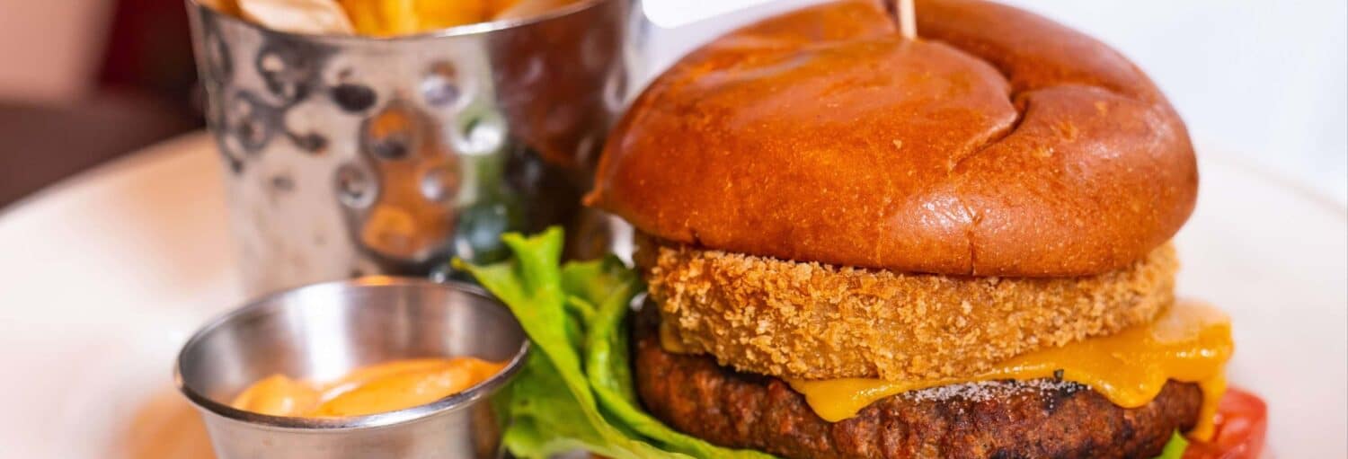 Um hambúrguer com alface, queijo e um hambúrguer frito, servido com batatas fritas em um copo de metal e um acompanhamento de molho. A foto é capa do post Hard Rock Cafe Porto sem filas.