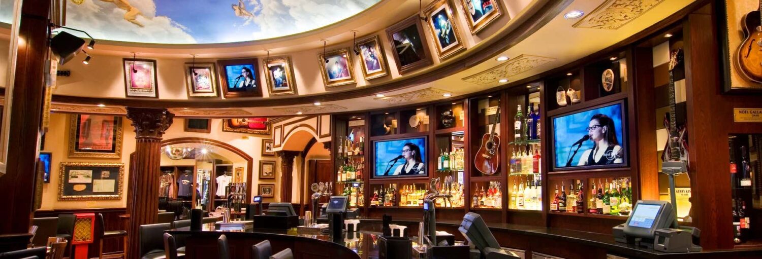 Interior do bar com quadros, guitarras e TVs acima de um balcão com garrafas e assentos; teto ornamentado com mural no Hard Rock Cafe Roma sem filas