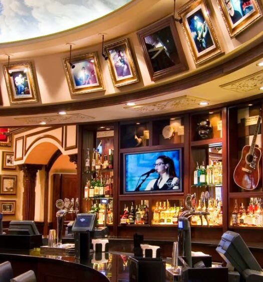 Interior do bar com quadros, guitarras e TVs acima de um balcão com garrafas e assentos; teto ornamentado com mural no Hard Rock Cafe Roma sem filas