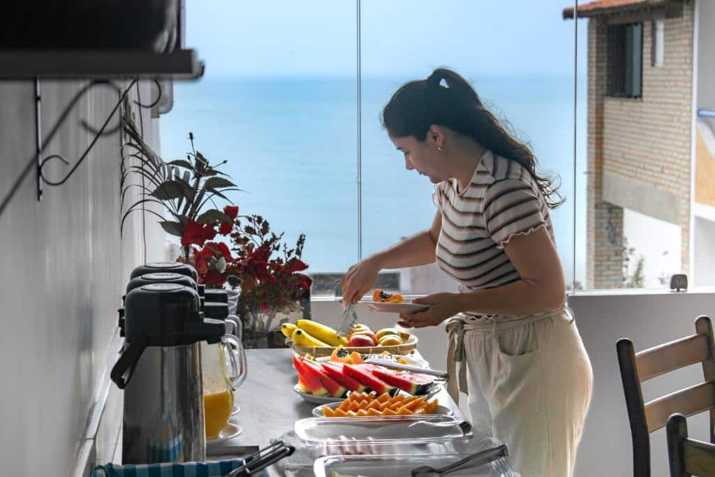 Uma mulher se serve de frutas em uma mesa de bufê com vários alimentos, disposta perto de uma janela com vista para o oceano.