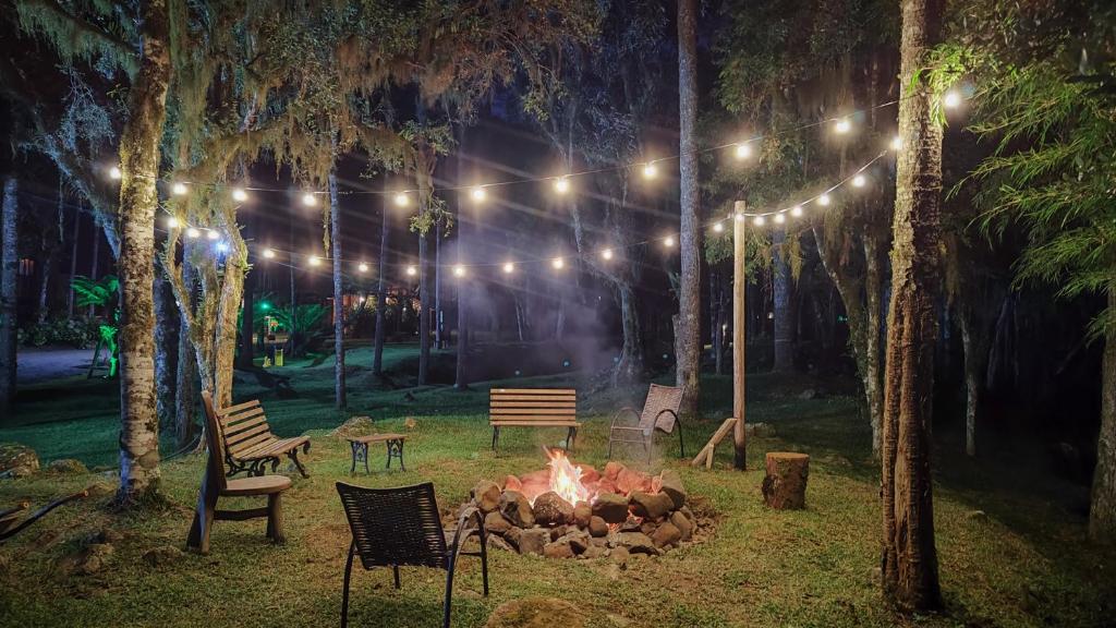 Uma fogueira com bancos e cadeiras é cercada por árvores e luzes de corda à noite em um ambiente externo gramado, na propriedade da Pousada Caminhos da Natureza.