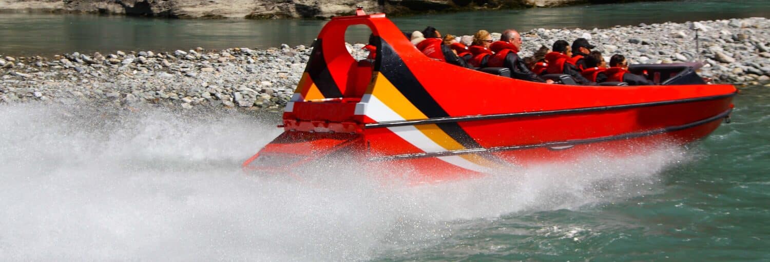 Um barco a jato vermelho com passageiros em coletes salva-vidas acelera na água, criando um borrifo perto de uma costa rochosa. A foto é capa do post sobre a Experiência Jet Boat no Porto.