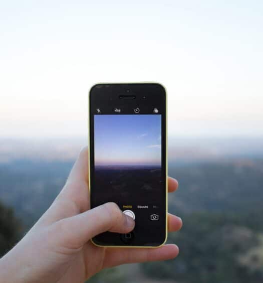 Uma mão segura um smartphone, tirando uma foto de uma paisagem com colinas e um céu claro ao fundo na Austrália. Ilustra o eSIM Oceania