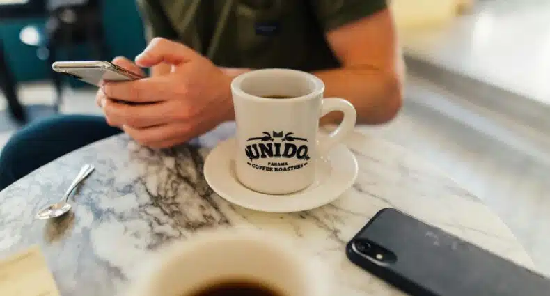 Pessoa usando um smartphone em uma mesa de mármore com uma caneca com a inscrição “Unido Coffee Roasters” e um telefone por perto.